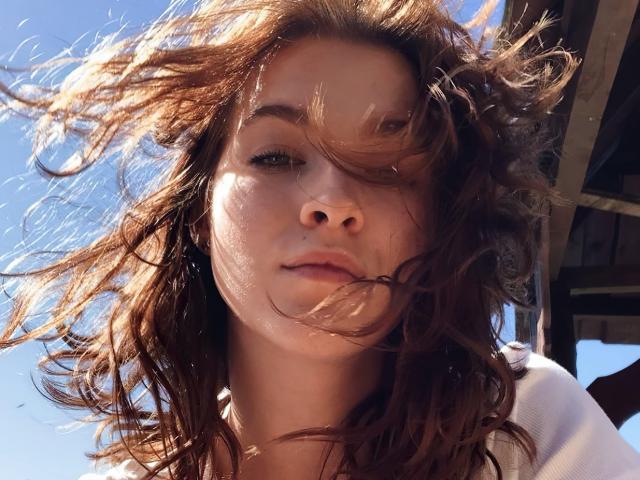 KenzieONEx's face with wavy brown hair blowing in the wind, blue sky, and sunlight. Her nose is centered, lips slightly parted, looking upwards. Soft shadows on her skin.