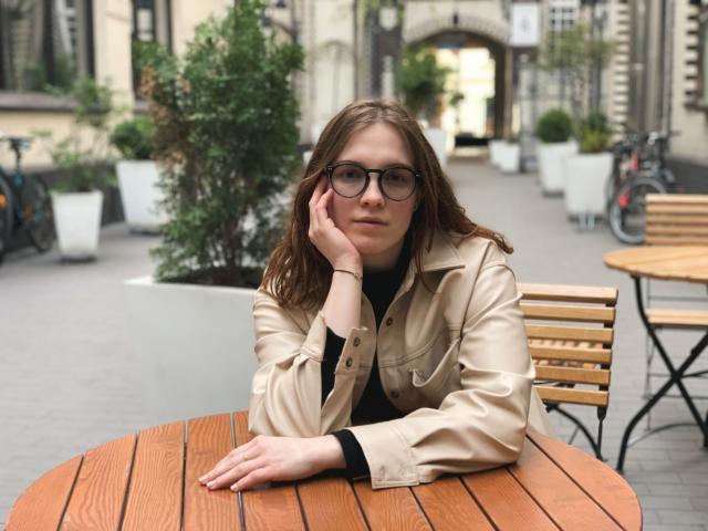 KenzieONEx with glasses, brown hair, and a beige jacket leans on a wooden table, hand on cheek, looking calm. Her tits are small, and she's cute.