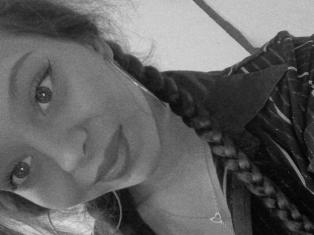 Autoportrait en noir et blanc de Bellefrance69, avec des cheveux tressés, portant une chemise rayée, des boucles d'oreilles cercles et un collier avec un petit pendentif en forme de cœur, regardant légèrement sur le côté..
