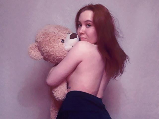 AnikaGlow with red hair, topless, hugging a fluffy teddy bear, wearing black pants, her back to the camera, looking over her shoulder.
