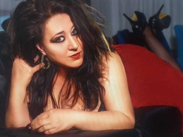 Una donna affascinante, formosa, con capelli scuri e ondulati, rossetto rosso e orecchini dorati, sdraiata su un cuscino rosso, che guarda verso l'alto con un'espressione seducente, e indossa tacchi alti neri..
