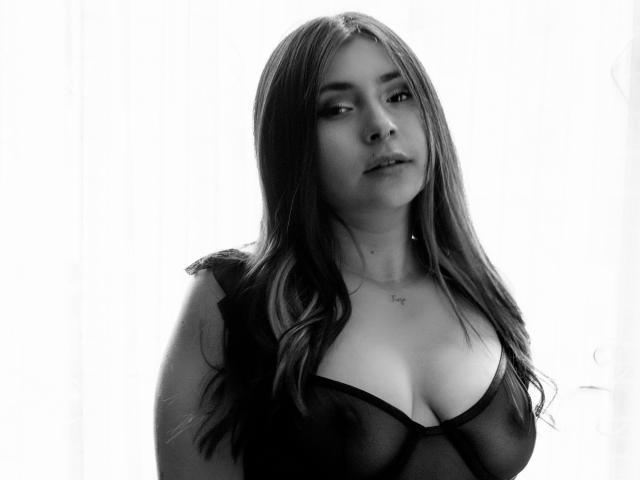 Black-and-white photo of MaryJaneHoe with long, wavy hair, wearing a sheer black top, showing her tits, and a subtle necklace against a bright, white background.