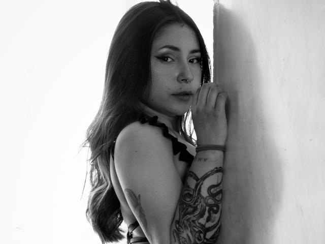 MaryJaneHoe, avec de longs cheveux noirs, un bras tatoué, un anneau au nez et un haut noir à volants, debout contre un mur, regardant en arrière, une main sur le mur, photo en noir et blanc..