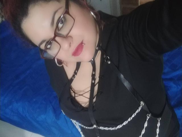 LatinoHardGirls with red hair, glasses, black outfit, chain details, and red lipstick, showing a slight smile, selfie with a blue background. Boobs visible, wearing a black top.