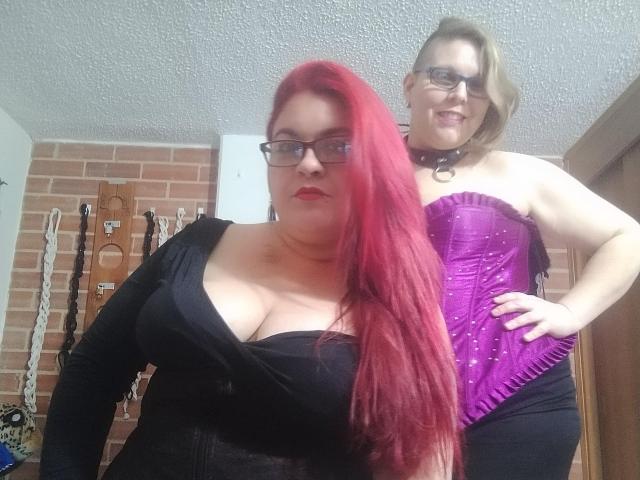 Two LatinoHardGirls, one with red hair and big tits in a black top, the other with blonde hair and a purple corset, standing in a room with BDSM gear.