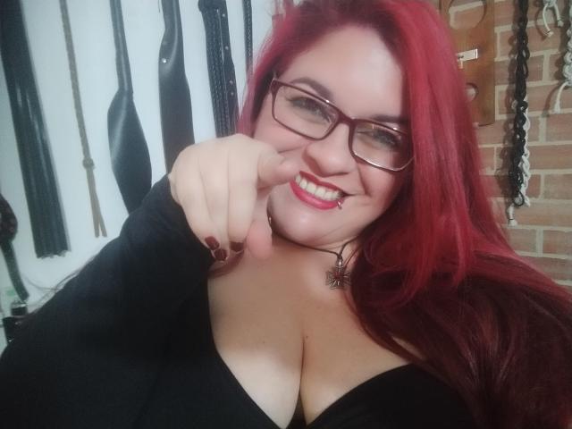 LatinoHardGirls with red hair, glasses, and black top, pointing at camera, smiling, showing big tits, red lipstick, and black choker with spider design.