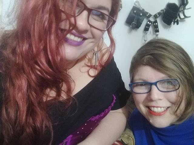 Two LatinoHardGirls with red and brown hair, glasses, purple lipstick, smiling, one in black and purple, other in blue, hanging BDSM gear in background.