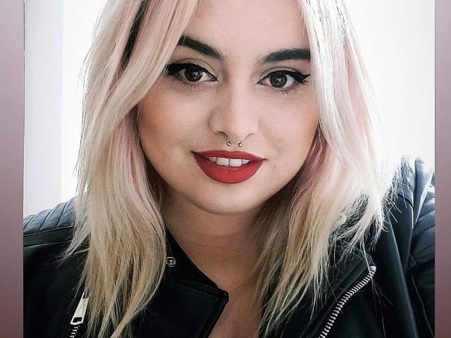 ChaudePourxToi ha i capelli biondi, l'eyeliner nero, il rossetto rosso, un piercing al naso e indossa una giacca di pelle nera, sorridendo alla fotocamera. Il suo seno non è visibile..