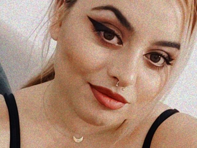 ChaudePourxToi has a nose ring, thick black eyeliner, orange lipstick, and a crescent moon necklace, with black straps visible on her chest. Blonde hair in a ponytail.
