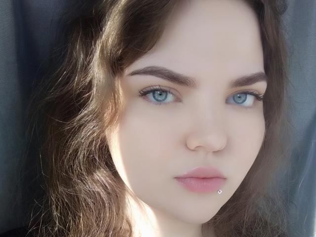 EishethZenunim tem pele clara, olhos azuis, cabelo castanho ondulado e um pequeno piercing de prata no lábio inferior. Ela está olhando diretamente para a câmera com uma expressão neutra..