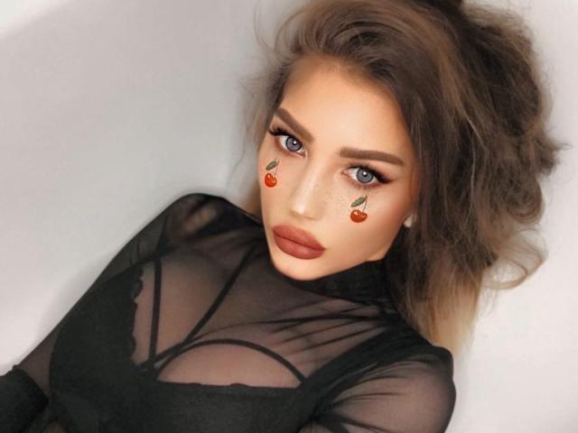 MARTINIK, een vrouw met kersenstickers op haar wangen, blauwe ogen en volle lippen, draagt een doorschijnend zwart topje, waardoor haar borsten zichtbaar zijn door de stof, en ligt plat..