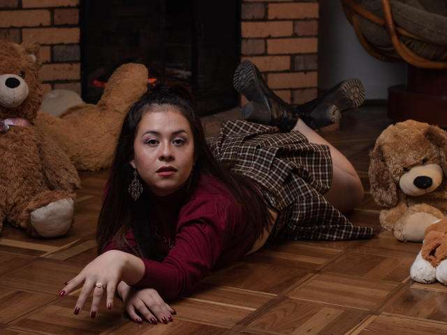 Katie Woolf, con lunghi capelli scuri, sdraiata sul pavimento di legno, indossa una camicia rossa, una gonna a quadri, calze nere e stivali, circondata da grandi orsi di peluche, guarda la fotocamera, con i glutei rivolti verso l'obiettivo..