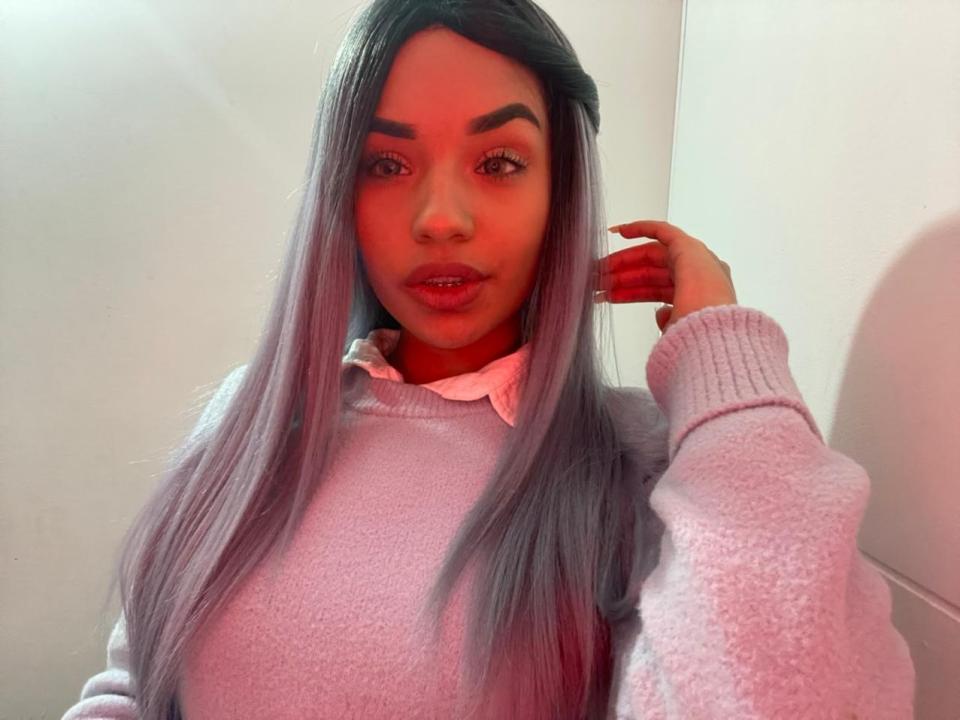 Keitlyn Reeves mit langem, graublauen Haar, trägt einen rosa, flauschigen Pullover, hat ihre Hand in der Nähe ihres Gesichts, ein rotes Licht auf ihrer Wange und ein leichtes Lächeln. Brüste sind unter dem Pullover sichtbar..