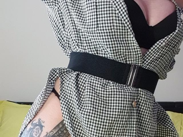 La robe à carreaux noirs et blancs de TonPlaisirDeReverBb, sa ceinture noire et son soutien-gorge noir, qui mettent en valeur sa poitrine et montrent un tatouage sur la cuisse droite, avec un oreiller jaune en arrière-plan..