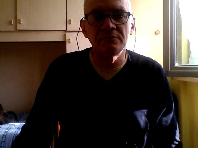 Man van middelbare leeftijd, kaal, met een bril, een zwart shirt en oordopjes, zittend in een zwak verlichte kamer. Er schijnt zonlicht van het raam aan de rechterkant, en hij kijkt licht geïrriteerd. SteelBg heeft een serieuze uitdrukking..