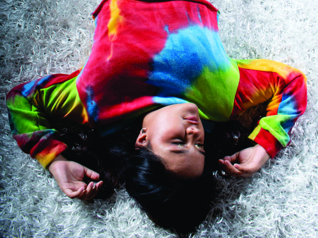 Il viso di KristalWade è capovolto, indossa una felpa colorata con motivi tie-dye, e i suoi capelli scuri sono sparsi su un tappeto bianco e soffice. Sembra rilassata, con le braccia alzate..