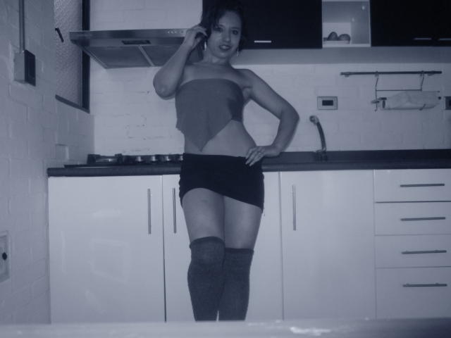 BellaLolita stands in a kitchen, talking on a phone, wearing a strapless top, black mini-skirt, and thigh-high socks, showing her ass and a flat belly.