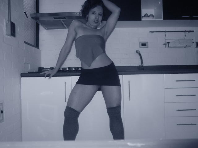 BellaLolita with bare tits, small ass, in a kitchen, wearing a strapless top, black mini-skirt, and thigh-high socks, standing with one hand on her head.
