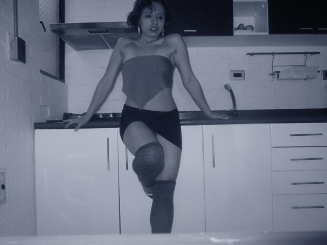 BellaLolita debout dans une cuisine, portant un haut sans bretelles, un short noir et un bas résille, avec une jambe levée, exposant sa vulve. Photo en noir et blanc, avec une teinte bleutée..