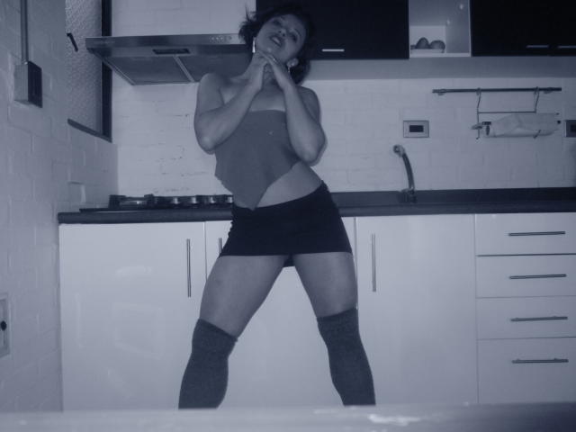 BellaLolita stands in a kitchen, wearing a tight tank top and black shorts, with her hands near her mouth, and thigh-high socks, looking sexy and confident.