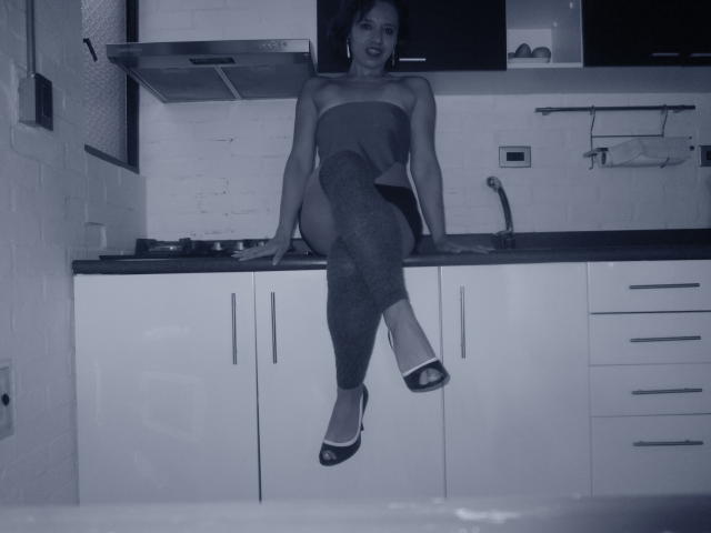 BellaLolita sitting on a kitchen counter, wearing a strapless top, dark leggings, and high heels, with her legs crossed, exposing her bare ass and small tits.
