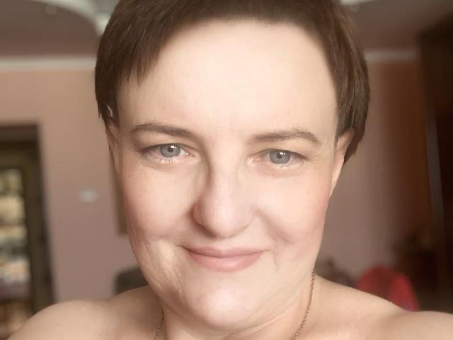 MargieEvans has short brown hair, light skin, blue eyes, and a slight smile. She's topless with a thin gold necklace, showing her bare shoulders.
