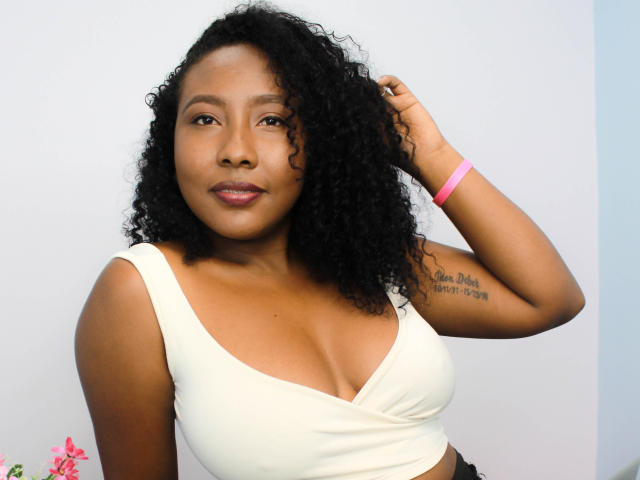 CaroldelaTorre, com cabelos pretos encaracolados, vestindo uma cropped branca que expõe seus seios, pulseira rosa, tatuagem no braço direito, pele marrom escura, lábios rosados, em pé contra um fundo branco..