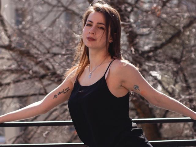SallyDesire has brown hair, a black tank top, small tits, tattoos on her right arm, and a necklace. She's leaning on a railing with a confident, relaxed expression.