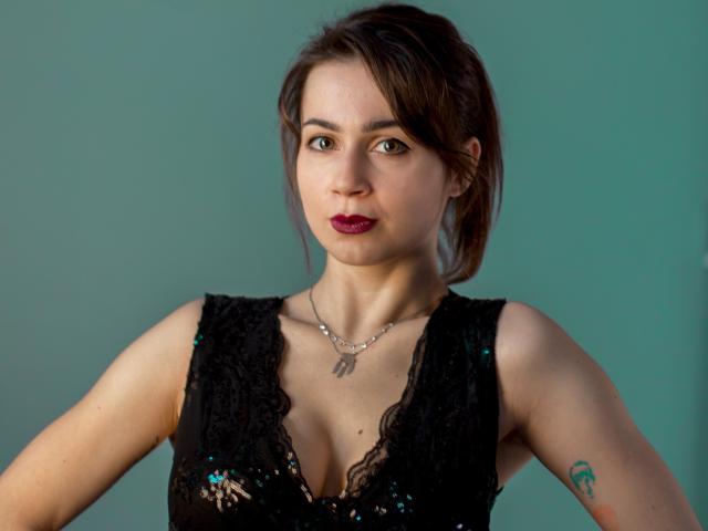 SallyDesire with brown hair, dark lipstick, black lace top, small tits, tattoo on right arm, necklace, green background, serious expression, medium skin, slight makeup, subtle eyeliner.