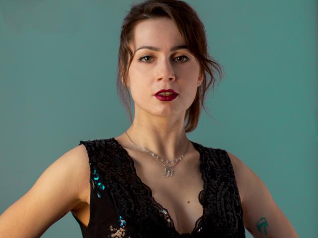 SallyDesire has brown hair, red lipstick, and wears a black lace dress with sequins, showing her tits, a silver necklace, and a tattoo on her right arm.