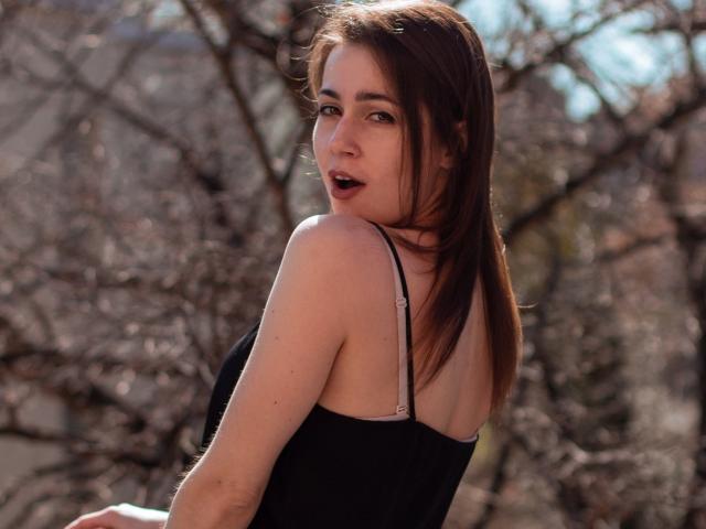SallyDesire tem cabelos castanho-médio, pele clara e olhos escuros, vestindo uma blusa preta com alças finas, olhando para trás com uma expressão ligeiramente surpresa e a boca aberta..