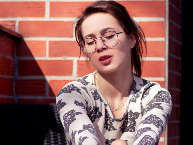 SallyDesire tem lábios vermelhos carnudos, óculos e uma blusa branca com estampas florais, com os braços cruzados e uma expressão relaxada. Ela está usando um colar dourado, em frente a uma parede de tijolos..