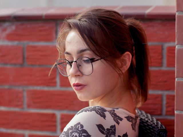 SallyDesire with brown ponytail, wearing glasses, and black-and-white butterfly patterned top, looking over shoulder, red brick wall in background. Her tits are small and she's cute.