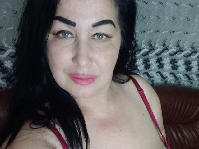LexyRose met zwart haar, groene ogen, dikke wenkbrauwen, roze lippenstift en een rode bh, kijkt naar de camera, met een lichte glimlach, heeft een lichte huid en zit op bruin leer..