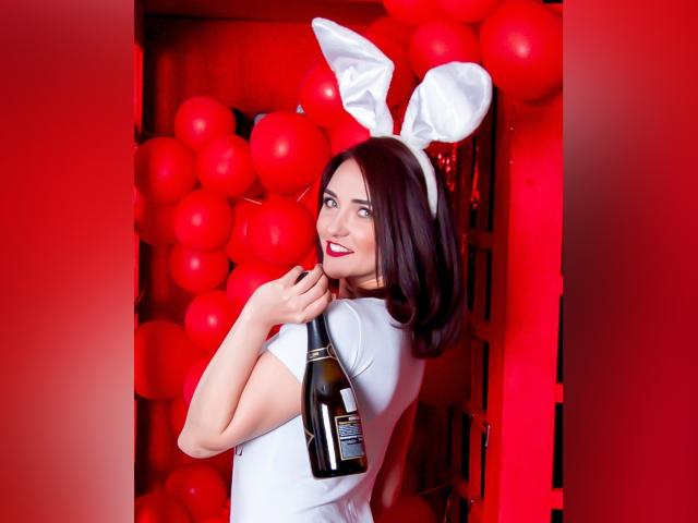 VeronicaLaLux con orejas de conejo blancas, sosteniendo una botella frente a una pared de globos rojos, sonriendo con lápiz labial rojo, cabello negro corto, camisa blanca y mirando hacia atrás..
