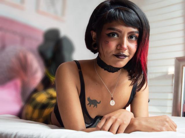 SerenaDeStark mit kurzen, schwarzen und roten Haaren, einem schwarzen Choker, dunklem Lippenstift, einem schwarzen BH und einem schwarzen Tattoo eines Hundes auf der Brust, liegt auf einem Bett und schaut in die Kamera..