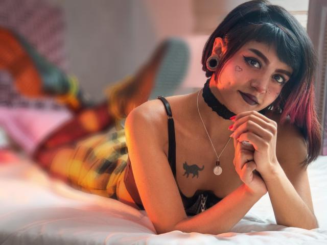 SerenaDeStark with black choker, dark lipstick, cat tattoo, and black bra, lying on bed, legs up, colorful socks, red and purple hair, ear gauges, and necklace, smiling.