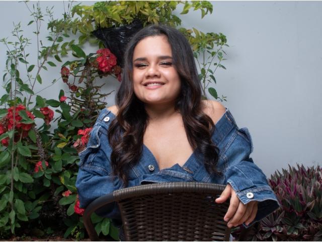 Kristal Wade, con lunghi capelli scuri, sorridente, indossa una camicia di jeans con una spalla scoperta, è seduta su una sedia di vimini, con le mani appoggiate allo schienale, circondata da fiori rossi e foglie verdi..