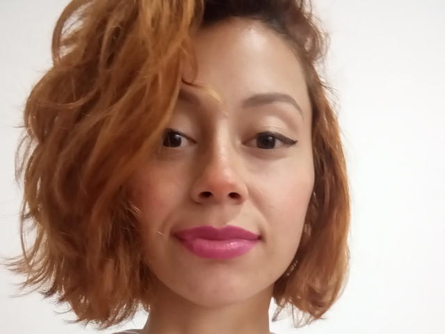 Eliza Jones tem cabelos castanhos claros ondulados, batom rosa e delineador preto, com um sorriso discreto e um tom de pele claro. Ela está olhando para o lado com olhos relaxados..