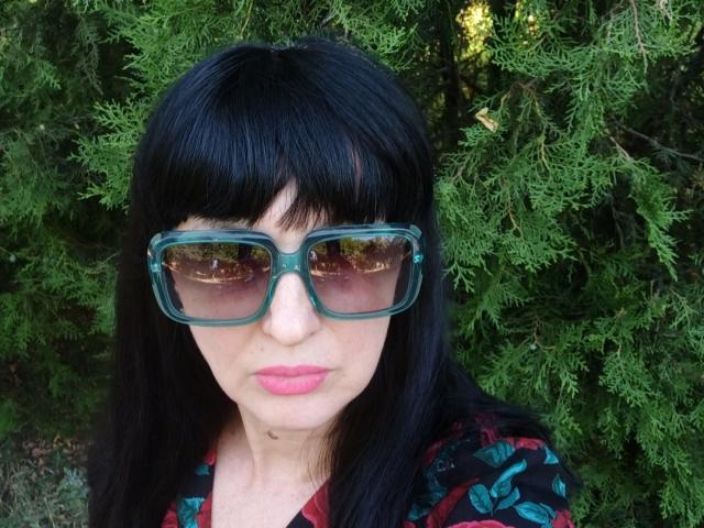 MelanyBanks with black hair, green-framed glasses, pink lipstick, and a dark patterned shirt, standing in front of green, leafy branches. Her nose is slightly upturned.
