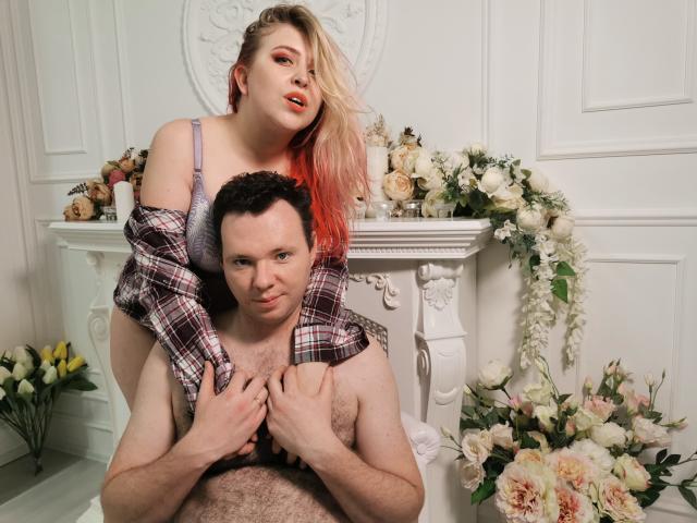 Genrikh Becca, con i capelli rosa e piccoli seni, in piedi, indossa un reggiseno grigio, sopra un uomo senza camicia, che si tiene il petto, circondata da fiori e pareti bianche..