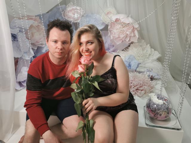 GenrikhBecca with pink rose, sitting beside GenrikhBecca in red-black striped sweater, black lace lingerie, holding rose, smiling, white curtain, floral decor, and crystal beads in background.