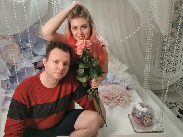 GenrikhBecca with pink hair, holding a rose, sits behind a chubby guy in a red and black striped shirt, looking at the camera with a smile. His hand's in her hair.