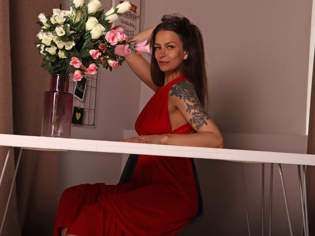 AlexiaBlack with long brown hair, red dress, tattoo on right arm, sitting on chair, hand in hair, white table, pink and white flower vase, smiling, brown curtains.