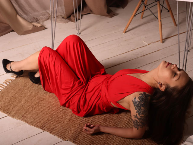 AlexiaBlack is lying on a burlap rug, wearing a red dress, black heels, and showing a tattoo on her right arm, with her head back and eyes closed.