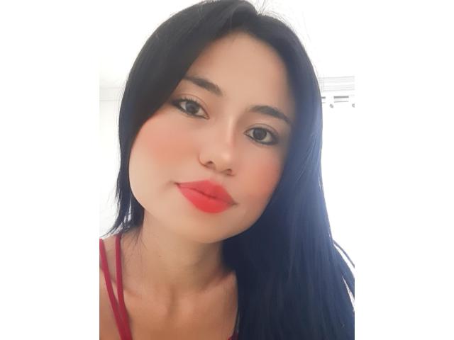 LolyFunX a les cheveux noirs, du rouge à lèvres, et une légère moue, elle porte un haut rouge, avec un visage clair et doux et des yeux marron, regardant la caméra..