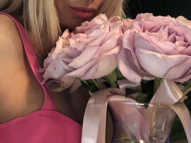 La camiseta rosa con abertura de AlisaAleksandr, su cabello rubio y su ramo de rosas rosas con cintas de seda, todo en un primer plano. Sus pechos parcialmente visibles..