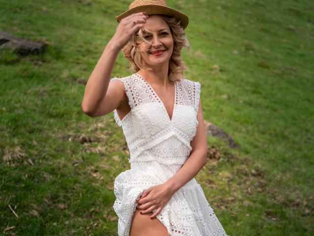 SoleildeNuit mit blonden Locken, lächelnd, trägt ein weißes Spitzenkleid, hält einen Strohhut und zeigt ein Bein im Gras. Ihre Hand liegt auf ihrem Genitalbereich, ihr Hintern ist bedeckt..
