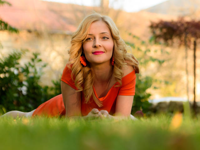 SoleildeNuit has blonde, curly hair, orange dress, and orange earrings, lying on green grass, smiling, with pink lipstick, and medium-sized tits, in a sunny outdoor setting.