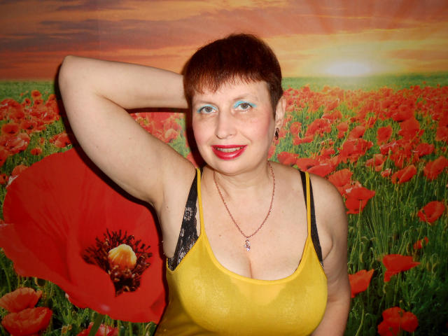 AirMagdalene met kort bruin haar, rode lippenstift, een geel tanktopje dat grote borsten laat zien, een zwarte kanten bh, arm achter het hoofd, een rode klaproos vasthoudend, lachend, met een rode ketting om..